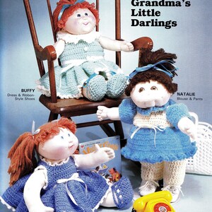 May include: Three Cabbage Patch Kids dolls dressed in crocheted outfits. The doll on the left is wearing a blue sundress and shoes. The doll in the middle is wearing a blue blouse and white pants. The doll on the right is wearing a green dress and shoes. The dolls are sitting on a wooden rocking chair and a white surface. The text "Grandma's Little Darlings" is printed above the dolls.