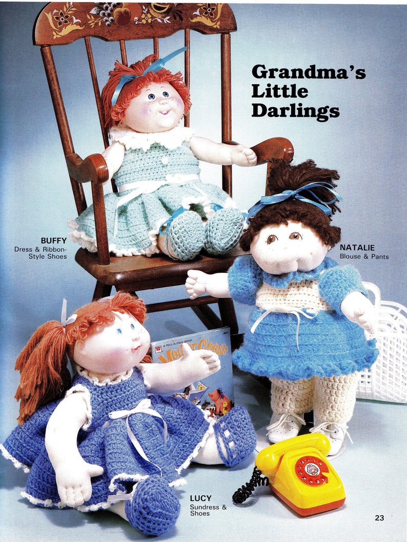 May include: Three Cabbage Patch Kids dolls dressed in crocheted outfits. The doll on the left is wearing a blue sundress and shoes. The doll in the middle is wearing a blue blouse and white pants. The doll on the right is wearing a green dress and shoes. The dolls are sitting on a wooden rocking chair and a white surface. The text "Grandma's Little Darlings" is printed above the dolls.