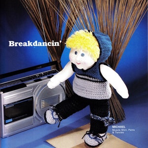 May include: A crocheted doll wearing a blue and gray muscle shirt, black pants, and blue and gray sneakers. The doll has blonde hair and blue eyes. The doll is breakdancing in front of a boombox. The text "Breakdancin'" is written in white letters on a blue background.