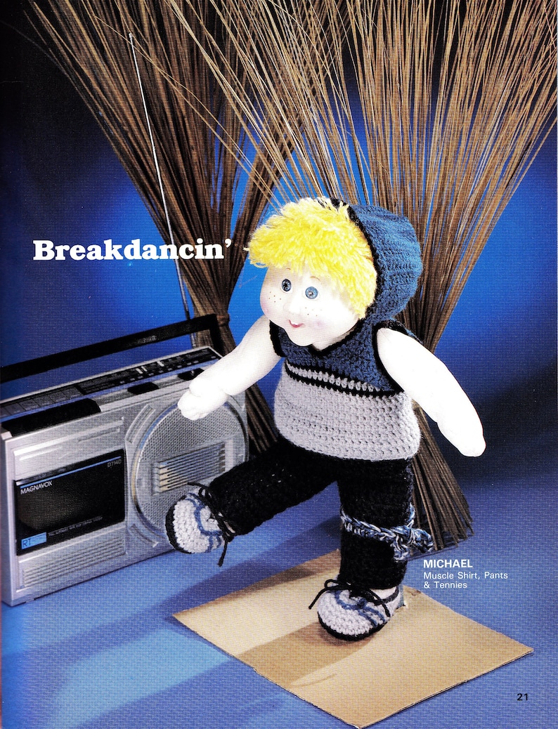 May include: A crocheted doll wearing a blue and gray muscle shirt, black pants, and blue and gray sneakers. The doll has blonde hair and blue eyes. The doll is breakdancing in front of a boombox. The text "Breakdancin'" is written in white letters on a blue background.