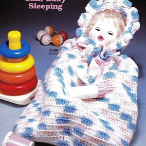 May include: A crocheted baby bunting with a pink and blue diamond pattern. The bunting has a hood with a pink and white crocheted trim. The bunting is lying on a blue surface with a baby doll inside. The doll has a pink and white crocheted hat and a white dress. The text "Shh, Baby Sleeping" is written above the bunting. The text "Susie Bunting" is written below the bunting.