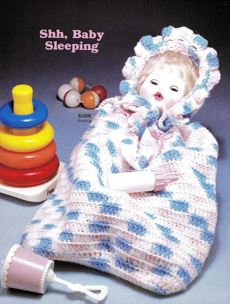 May include: A crocheted baby bunting with a pink and blue diamond pattern. The bunting has a hood with a pink and white crocheted trim. The bunting is lying on a blue surface with a baby doll inside. The doll has a pink and white crocheted hat and a white dress. The text "Shh, Baby Sleeping" is written above the bunting. The text "Susie Bunting" is written below the bunting.