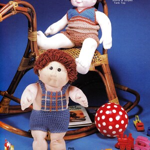 May include: Two Cabbage Patch Kids dolls dressed in crocheted outfits. The doll on the left is wearing a blue and orange overalls and the doll on the right is wearing a striped tank top and shorts. The dolls are sitting on a rocking chair and playing with toys.