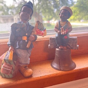 May include: Two figurines of young boys, one in a blue uniform holding an American flag and a bag, the other sitting on a bench holding a smaller American flag, both with brown skin and black hair, on a wooden surface.