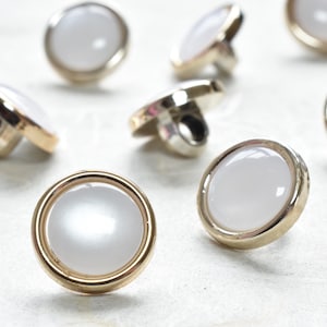 May include: A collection of pearlized white buttons with gold-colored rims and shanks. The buttons are scattered across a light-colored surface, showcasing their reflective quality and design.
