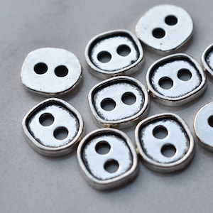 Puede incluir: Diez botones plateados con dos agujeros cada uno. Los botones tienen una forma irregular y una superficie ligeramente texturizada.