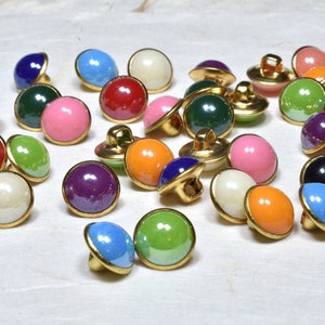 Puede incluir: Surtido de botones coloridos con bordes de metal dorado. Los botones son de varios colores, incluyendo rojo, verde, azul, rosa, naranja, morado y blanco.