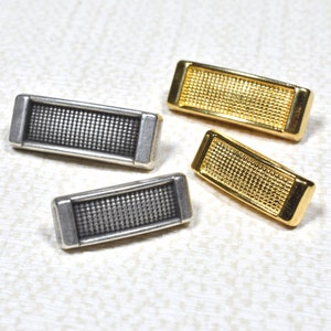 Op de afbeelding: Drie rechthoekige metalen versieringen met een getextureerd oppervlak. Twee zijn goud en één is zilver.