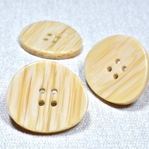 May include: Three oval-shaped buttons with a wood grain pattern. The buttons are beige with a light brown wood grain pattern. The buttons have different numbers of holes. One button has two holes, one has four holes, and one has five holes.