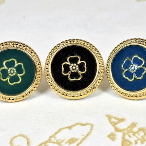 Puede incluir: Tres botones redondos con bordes dorados y un diseño de flor en el centro. Los botones son verde, negro y azul.