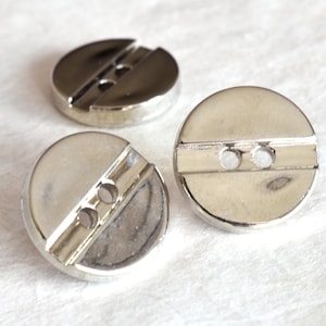 Puede incluir: Tres botones de metal plateado con una línea horizontal en relieve y dos agujeros.