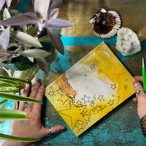 May include: A white canvas with a yellow and gold abstract background. The canvas is decorated with black outlined stars of different sizes. The largest star is in the center of the canvas and is surrounded by smaller stars.