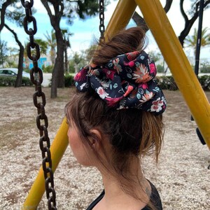 Può includere: Una donna con i capelli castani raccolti in uno chignon con un elastico per capelli con motivo floreale. L'elastico è nero con fiori rosa, arancioni e bianchi.