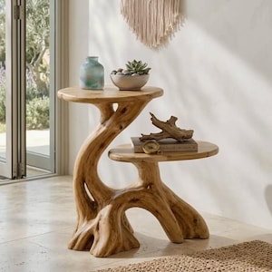 May include: A two-tiered side table crafted from light-toned natural wood with a gnarled, root-like base. The top tier holds a blue-tinted glass vase and a small bowl with succulents, while the lower tier supports a book and a piece of driftwood.