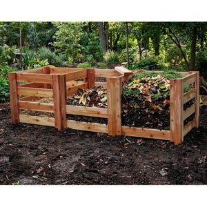 May include: A three-section wooden compost bin constructed from horizontal cedar slats. The structure features open-slat walls and contains organic waste, including dry straw, dark soil, and various food scraps like banana peels and avocado skins.