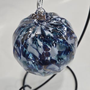 Peut inclure: Une boule de Noël en verre bleu et blanc avec une finition métallisée argentée. La boule a une surface texturée et porte l'année "2024" sur une étiquette argentée.