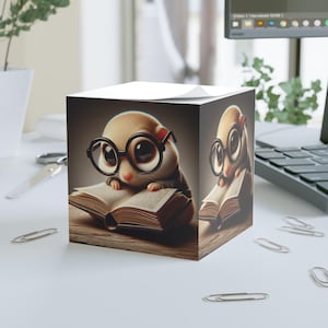 May include: A cube-shaped notepad with a cartoon illustration of a small rodent wearing glasses and reading a book. The image has a warm colour palette and a whimsical theme, suitable for office or study use.