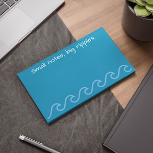 Può includere: Un blocco note blu turchese con la scritta "Small notes, big ripples" in corsivo bianco. Il blocco note presenta un motivo a onde lungo il bordo inferiore. È appoggiato su una superficie grigia, con un laptop, una penna e un taccuino sullo sfondo.