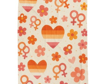 Op de afbeelding: Een crèmekleurige deken met een herhaald patroon van oranje en roze harten, bloemen en vrouwelijke symbolen. De harten hebben oranje en roze strepen. De bloemen en symbolen zijn in tinten oranje en roze.