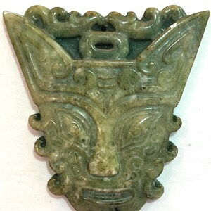 Op de afbeelding: Een gebeeldhouwde jade hanger in groentinten, met een gestileerd gezicht met prominente ogen, een neus en een mond. De hanger heeft een gedetailleerde, sierlijke hoofdtooi en een symmetrisch ontwerp. De jade heeft een gepolijste afwerking.