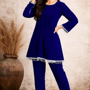 May include: A two-piece, royal blue outfit. The top has long sleeves with white fringe at the cuffs, and the hemline is also trimmed with white fringe. The matching pants are a straight-leg style. The outfit is worn with white heels.