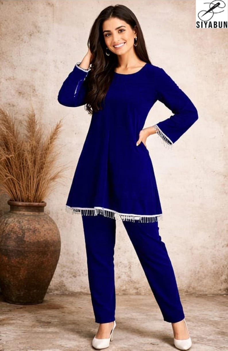 May include: A two-piece, royal blue outfit. The top has long sleeves with white fringe at the cuffs, and the hemline is also trimmed with white fringe. The matching pants are a straight-leg style. The outfit is worn with white heels.