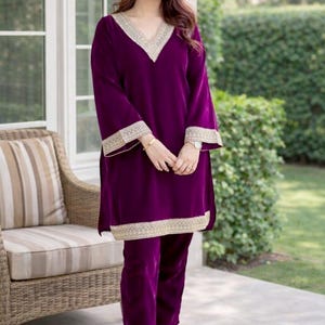 Könnte beinhalten: Ein tiefviolettes Samt-Outfit, bestehend aus einer langärmeligen Tunika mit V-Ausschnitt und passender Hose. Die Tunika und die Ärmel sind mit goldfarbener Stickerei verziert. Das Outfit wird mit goldfarbenen Schuhen getragen. Das Markenlogo "SIYABUN" ist sichtbar.