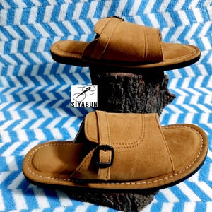 May include: Two brown suede sandals with a buckle closure are displayed on a wooden surface against a blue and white patterned background. The sandals have a stitched design and a dark sole. The brand name "SIYABUN" is visible.