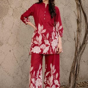 May include: A deep red two-piece set consisting of a long-sleeved tunic with a mandarin collar and button placket, paired with wide-leg trousers. Both pieces feature a large-scale, cream-colored botanical floral print along the lower sections.