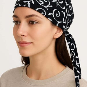 May include: A black headscarf featuring a white, swirling floral design. The head covering is tied at the back, with the ends trailing down. The Siyabun logo is visible in the top right. The person in the image is wearing a beige top.