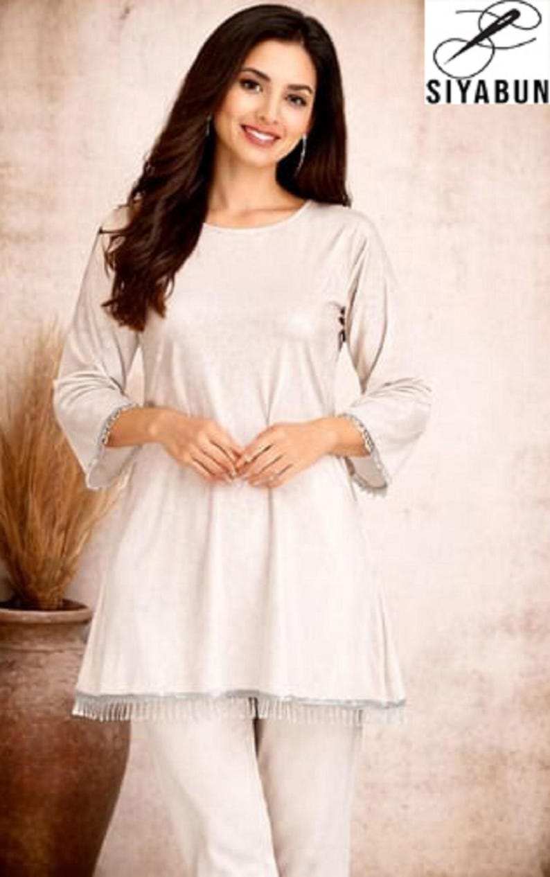May include: A light beige two-piece outfit. The top has long sleeves with a decorative trim at the cuffs and hem. The outfit is made of a soft, flowing fabric. The logo "SIYABUN" is in the upper right corner.