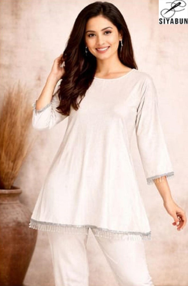 May include: A woman wearing a white tunic and matching pants. The tunic has three-quarter sleeves and a decorative trim. The outfit is simple and elegant, suitable for casual or semi-formal occasions. The brand logo "SIYABUN" is visible in the top right corner.