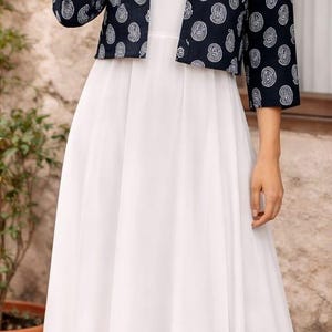 May include: A woman wearing a white dress and a navy blue jacket with a white paisley pattern. The dress is floor-length, and the jacket is cropped. The image is taken outdoors.