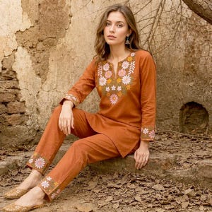May include: A rust-coloured long-sleeve tunic and matching trousers set featuring a floral embroidery pattern in white, pink, and green around the neckline and on the sleeve cuffs and pant hems. The outfit is made of a solid-coloured fabric.