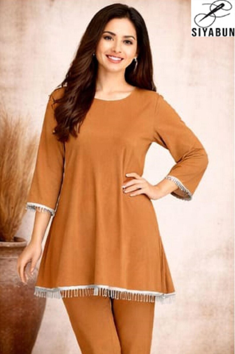 May include: A two-piece outfit in a warm brown hue. The top features three-quarter sleeves and a flared hemline, both trimmed with white fringe. The matching pants complete the ensemble. The brand logo "SIYABUN" is visible in the upper right corner.
