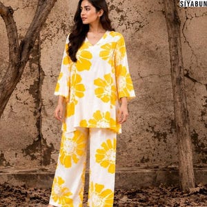 May include: A two-piece outfit consisting of a white tunic with a V-neckline and three-quarter length bell sleeves, paired with matching wide-leg trousers. Both pieces feature a bold, large-scale yellow floral print. The logo SIYABUN is visible.
