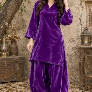 May include: A deep purple velvet tunic and matching loose-fitting trousers. The tunic features a V-neckline and long, wide sleeves. The trousers have a gathered, voluminous silhouette. The fabric has a soft, reflective sheen throughout the set.