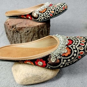 May include: A pair of black embroidered flat shoes with silver embellishments. The shoes have a pointed toe and a low heel. The embroidery features floral and geometric patterns in red, orange, and white.
