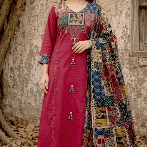 May include: A deep red long-sleeved kurta with a V-neckline, featuring a patchwork-style patterned yoke and cuffs. The garment includes three decorative tassels down the front and is paired with matching wide-leg trousers and a patterned shawl.