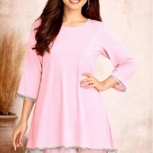 May include: A woman wearing a light pink tunic and matching pants. The tunic has three-quarter sleeves and a silver fringe trim. The outfit is from the brand SIYABUN, as indicated by the logo in the upper right corner.