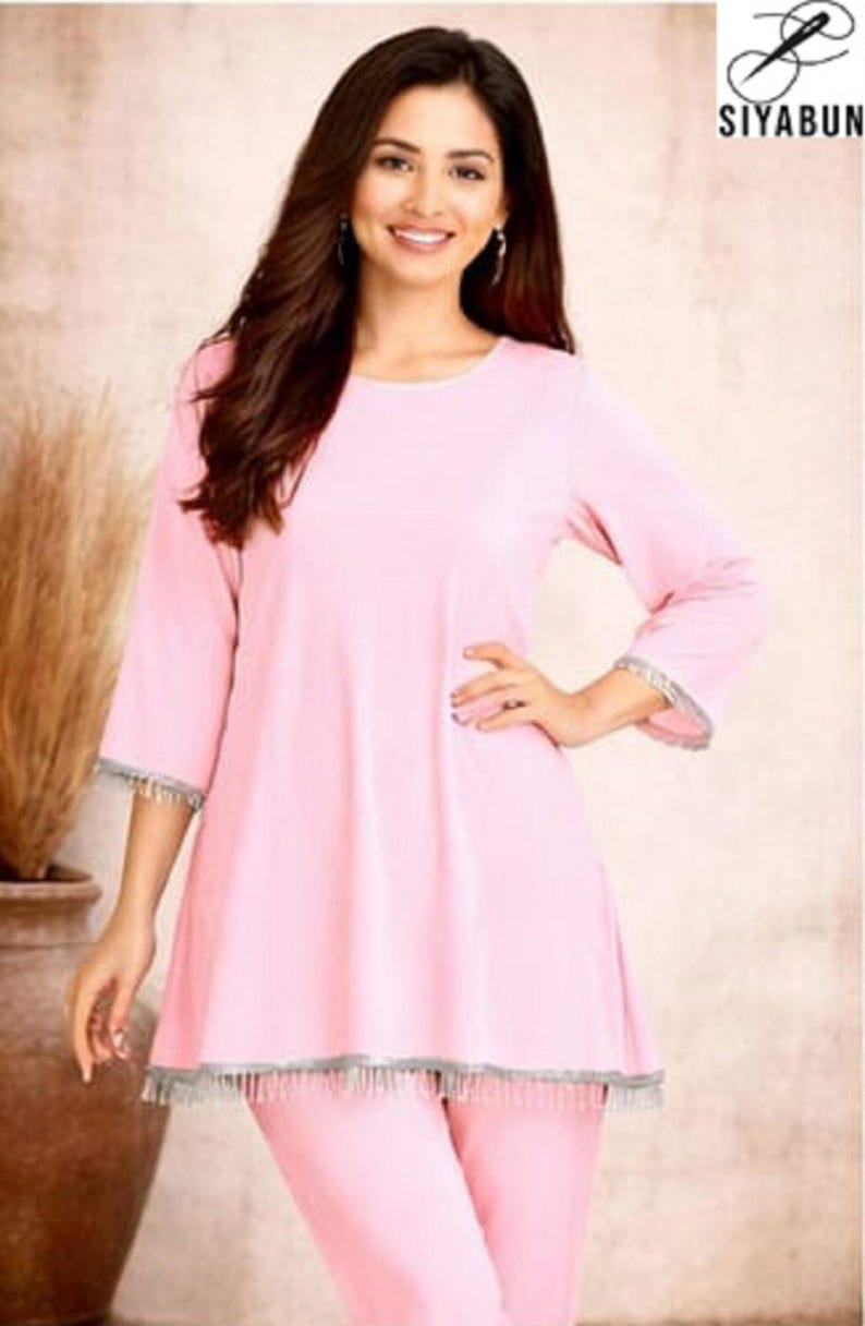 May include: A woman wearing a light pink tunic and matching pants. The tunic has three-quarter sleeves and a silver fringe trim. The outfit is from the brand SIYABUN, as indicated by the logo in the upper right corner.