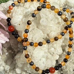 Op de afbeelding: Drie armbanden met kralen in aardetinten, waaronder oranje, bruin, zwart en grijs. De armbanden liggen op een wit, kristallijn oppervlak. De kralen variëren in grootte en textuur. Op de achtergrond is een roze bloem te zien.