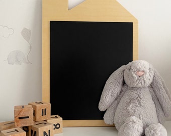 May include: A light-colored wooden chalkboard shaped like a house with a black rectangular writing surface. Beside the board sits a soft, plush grey rabbit toy with long ears and a pink nose, and several wooden blocks with black letters and numbers.