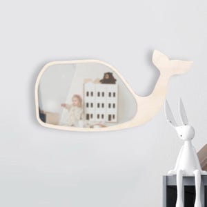 May include: A wall-mounted mirror shaped like a whale, featuring a light-colored wooden frame. The reflective surface shows a blurred interior scene with a small toy house and a child. The whale design includes a distinct tail fin on the right side.