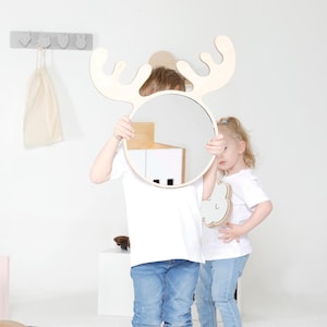 May include: A circular wall mirror featuring a light-colored wooden frame shaped like moose antlers. The mirror is held up by a person, obscuring their face. The frame has a smooth, natural wood finish and a simple, clean design for wall mounting.