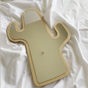 May include: A cactus-shaped wall mirror featuring a light-colored wooden frame with a smooth finish. The reflective glass surface follows the contour of the wooden base, which has two side branches extending outward from the central vertical body.
