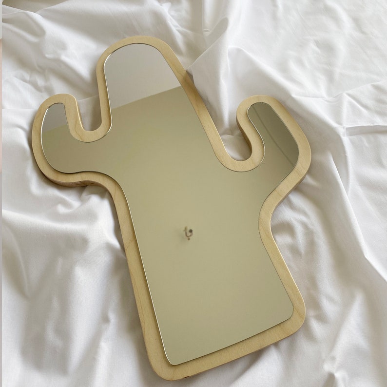 May include: A cactus-shaped wall mirror featuring a light-colored wooden frame with a smooth finish. The reflective glass surface follows the contour of the wooden base, which has two side branches extending outward from the central vertical body.