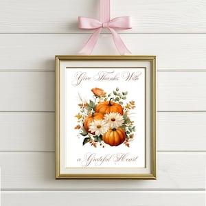 May include: A framed print with a gold border, featuring a watercolor illustration of pumpkins, daisies, and foliage. The text reads "Give Thanks With a Grateful Heart." A pink ribbon is tied at the top for hanging.