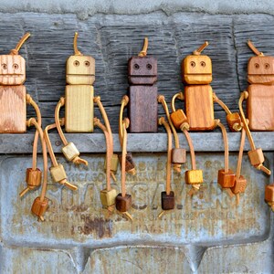 May include: Five wooden robot figurines with leather straps and wooden beads. The robots are different shades of brown and are arranged in a row on a metal surface.