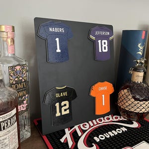 Pode incluir: Um painel de exibição preto com quatro camisolas desportivas em miniatura com nomes e números de jogadores: um Nabers 1 azul escuro, um Jefferson 18 roxo, um Olave 12 preto e um Chase 1 laranja. O painel está posicionado entre várias garrafas.
